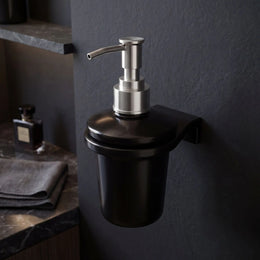 Black soap dispenser with silver pump on a dark wall in a bathroom setting.